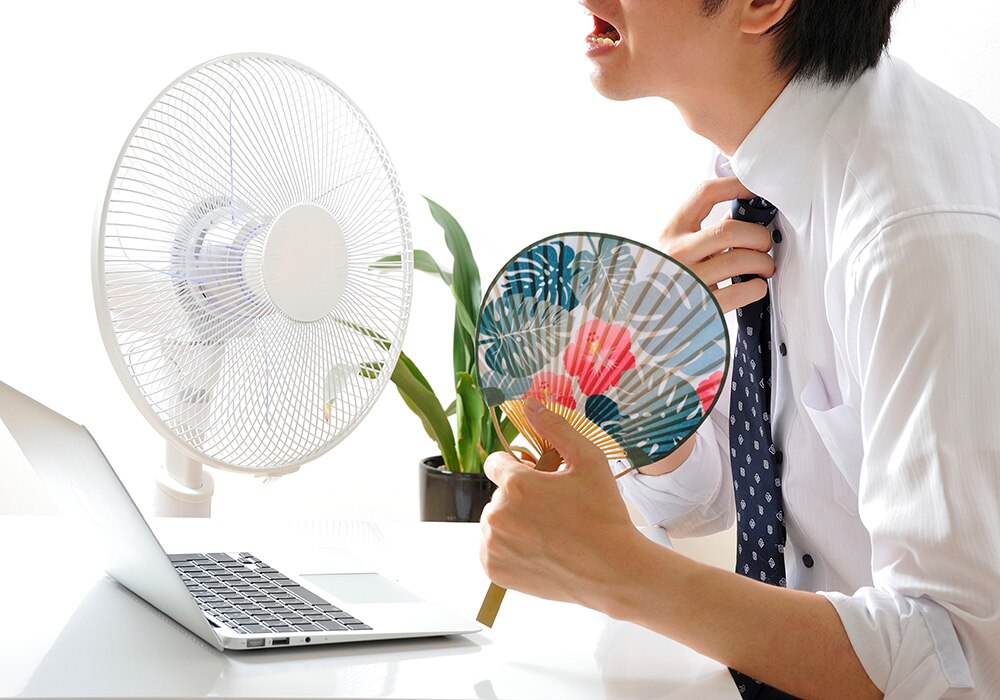 暑いオフィスで扇風機とうちわを使っているビジネスパーソン